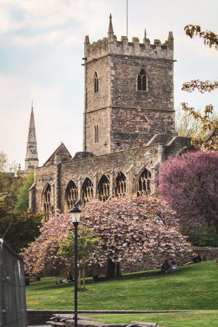 A historic stone church with tall, narrow arched windows and crenellated battlements at the top of its square tower is set against a partly cloudy sky. In the foreground, a well-maintained garden feat