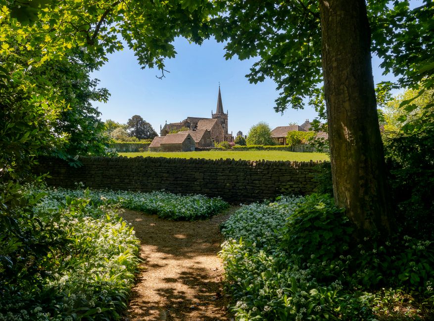 A historic stone church with tall, narrow arched windows and crenellated battlements at the top of its square tower is set against a partly cloudy sky. In the foreground, a well-maintained garden feat